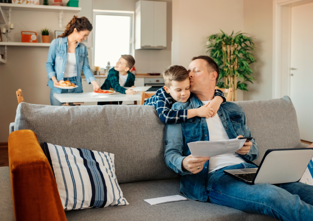 A mother and son preparing lunch at the dinning table, a son leaning over the back of a couch while the father is working their